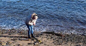 A woman beachcombing on the coastline on a pebbled beach next to the sea